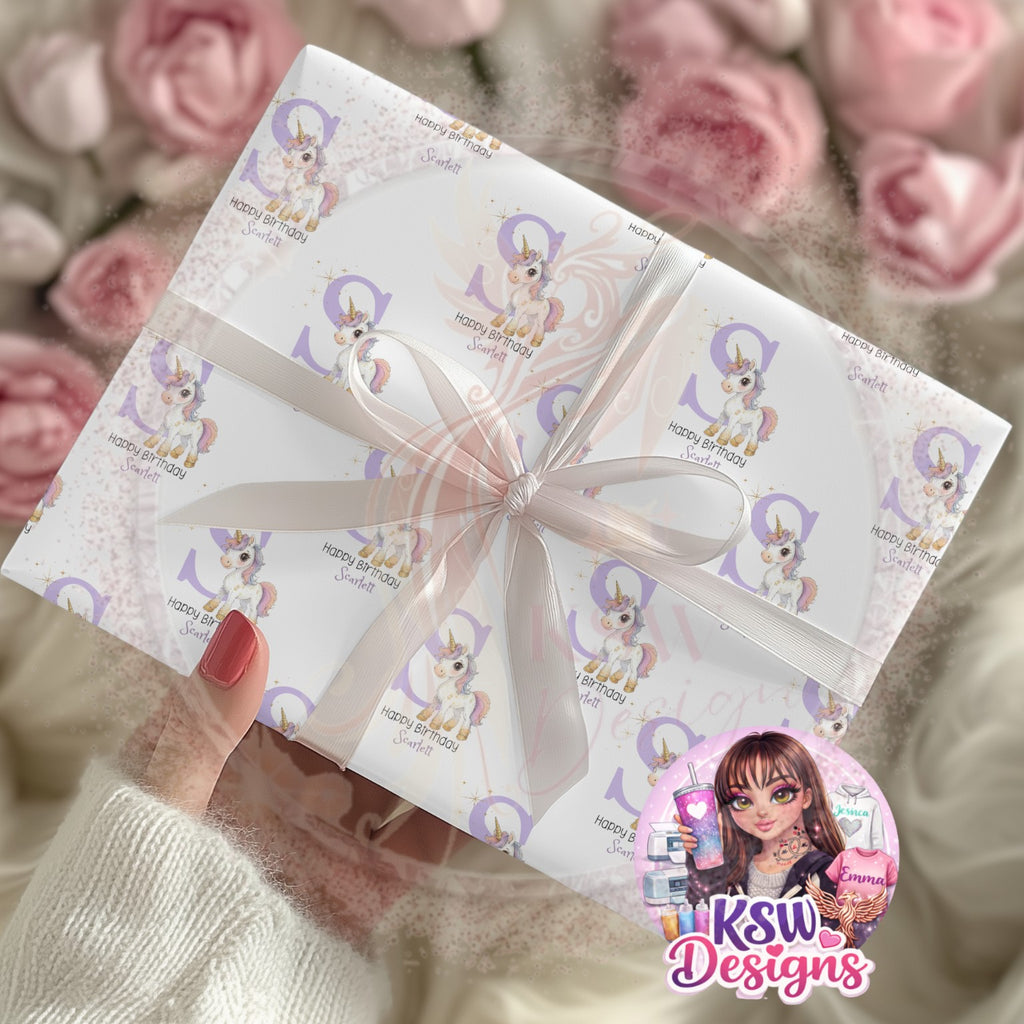 Personalised unicorn birthday wrapping paper featuring a pastel unicorn, large lilac letter S, golden sparkles and Happy Birthday Scarlett wording, shown wrapped around a gift with a white ribbon